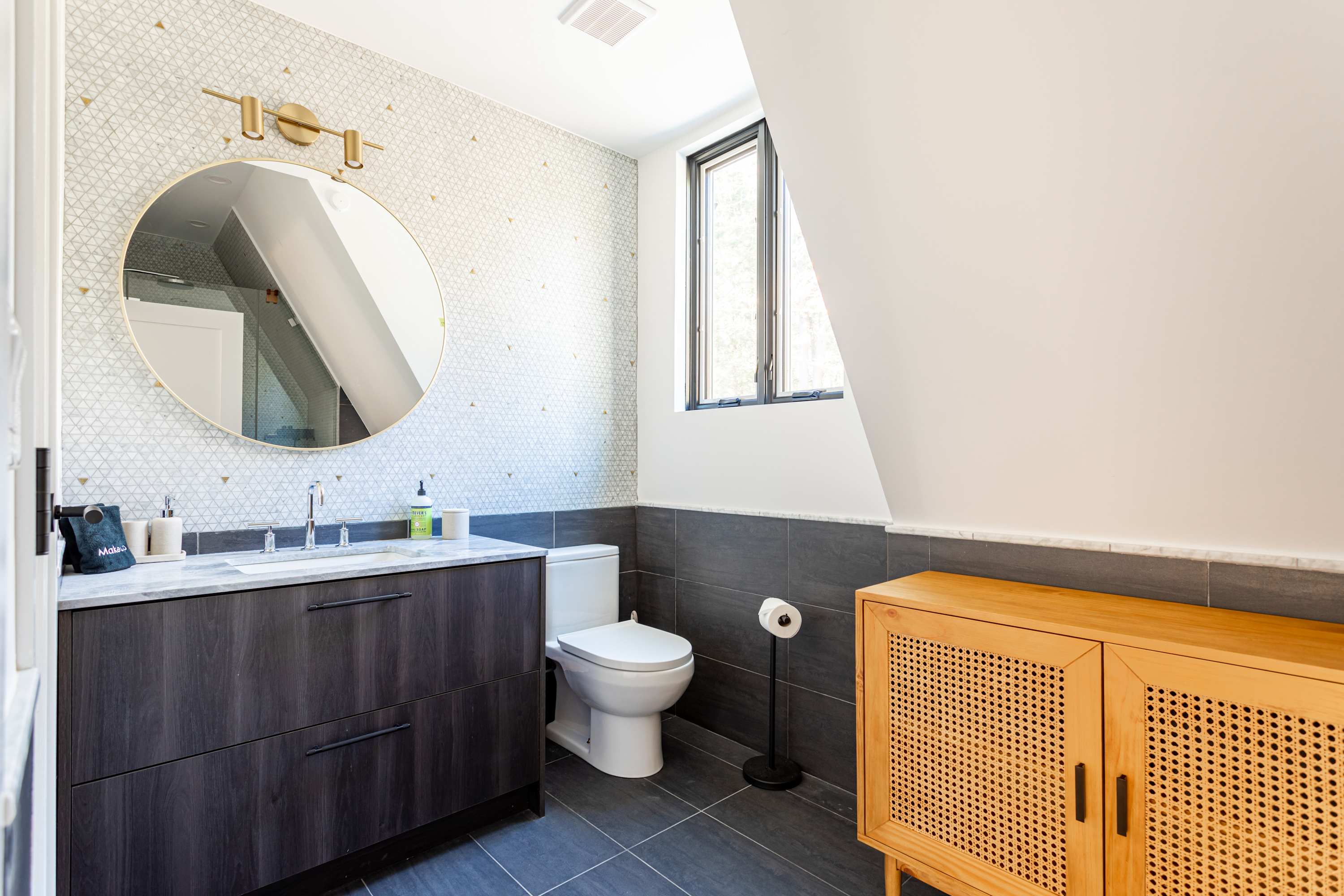 A modern bathroom featuring a dark wood vanity with a white marble countertop and an undermount sink. A large round mirror with a gold frame hangs above, and the wall is adorned with geometric patterned tiles.