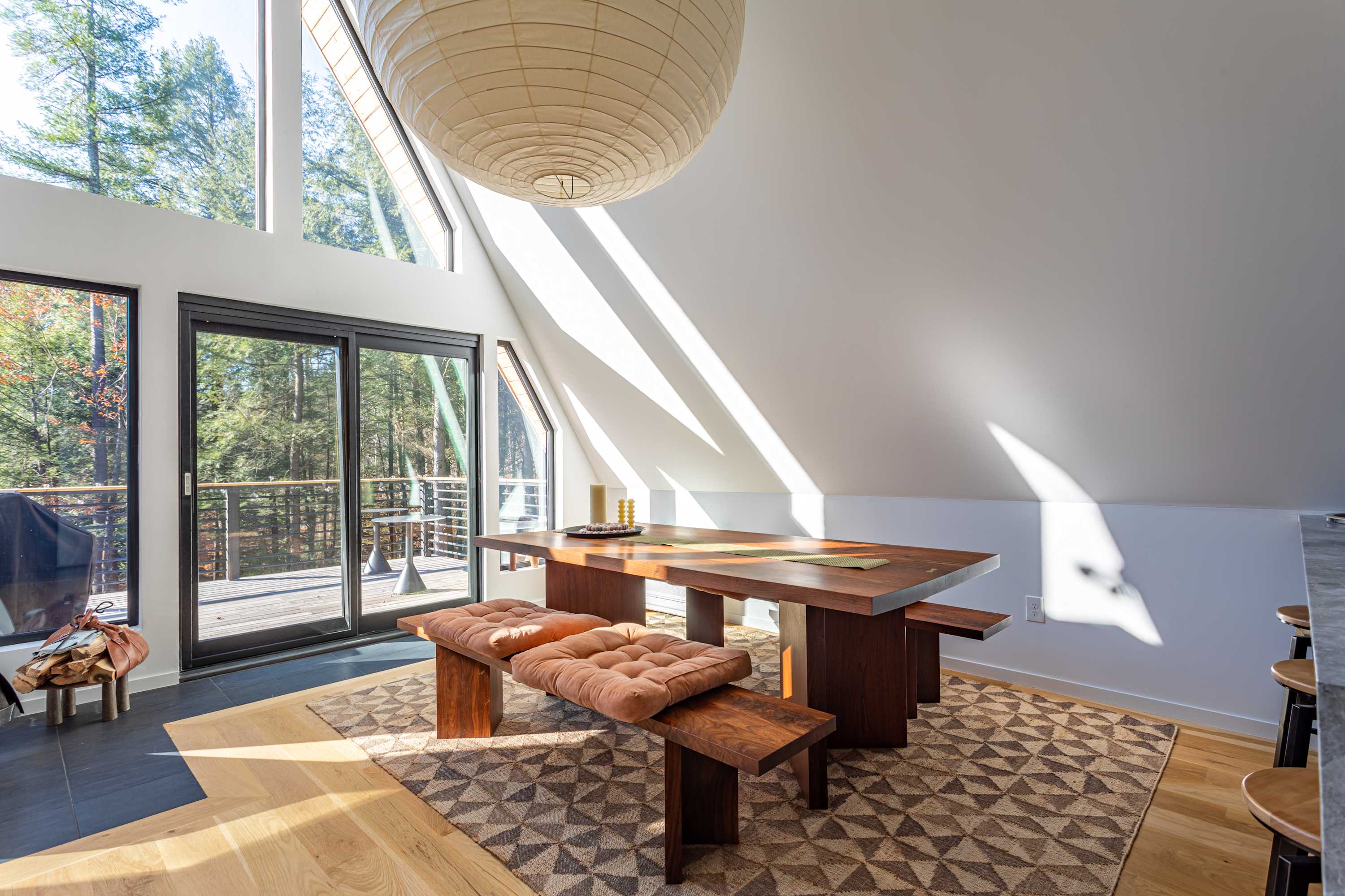 A close-up of the dining area features a dark wooden table with matching benches, adorned with orange cushions, set on a geometric patterned rug. Large sliding glass doors open to an outdoor deck with forest views.