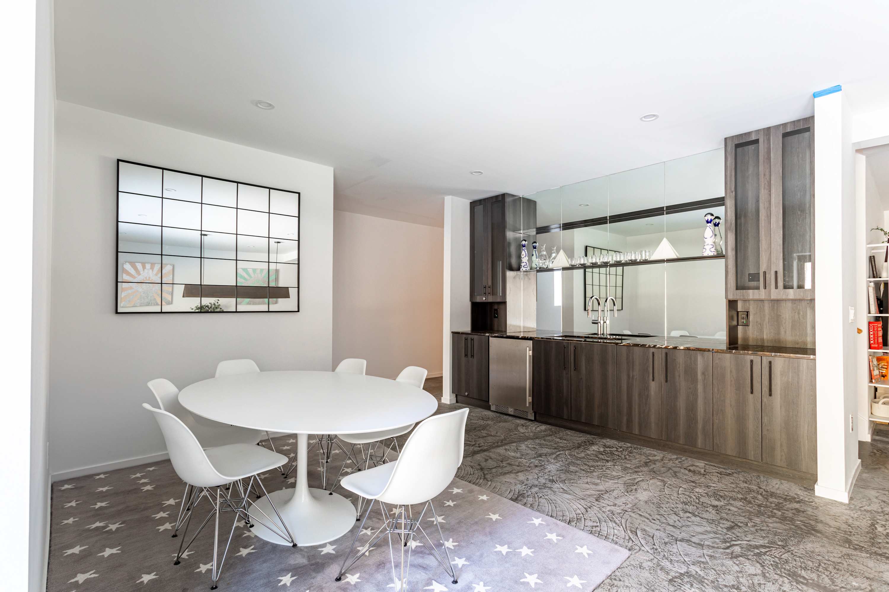 A modern dining area features a round white table with six white chairs on a gray rug with white stars, adjacent to a wet bar with dark wood cabinetry, a mirrored backsplash, and stainless steel appliances.