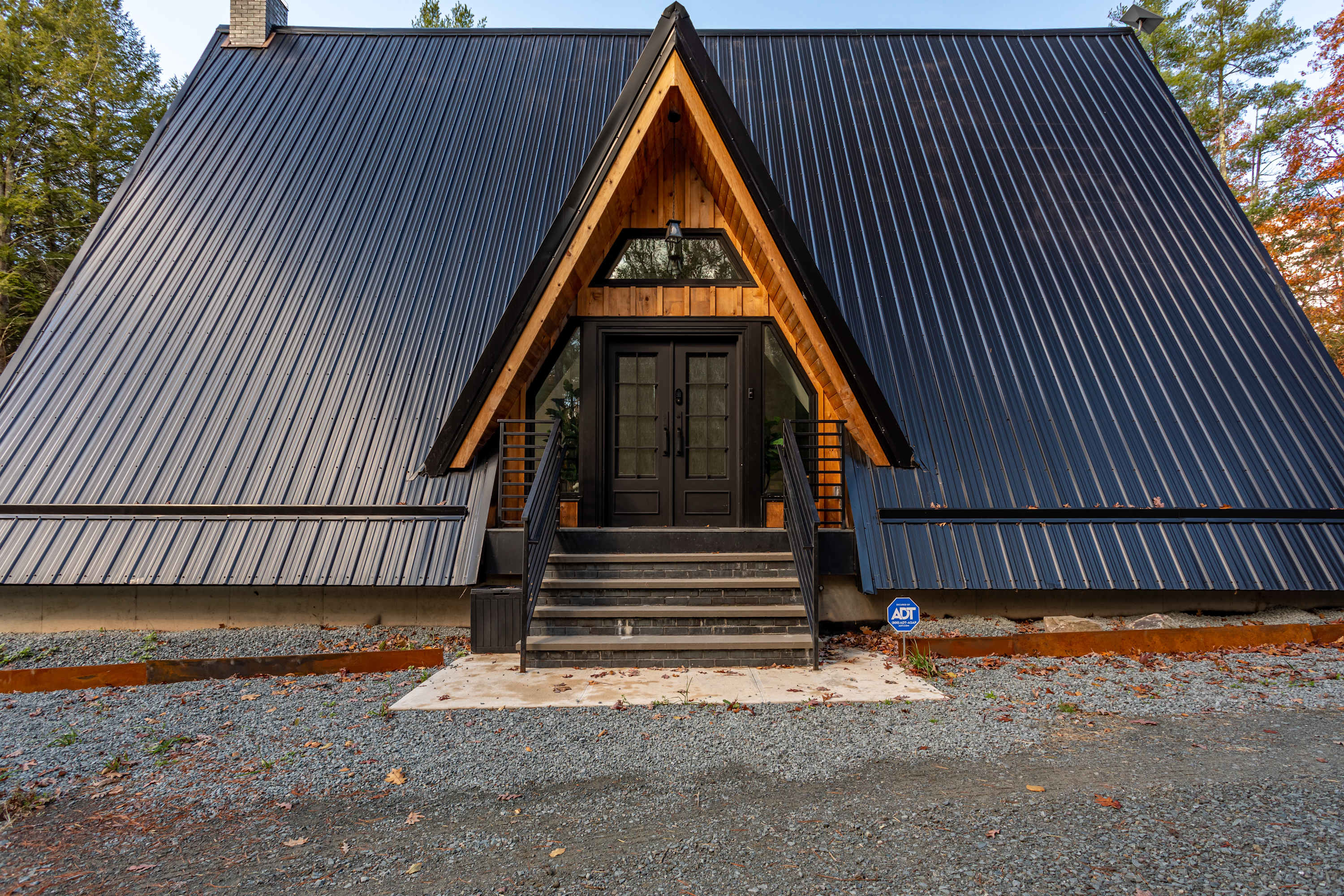 The front exterior of a modern A-frame house features a dark metal roof, natural wood accents around the entrance, and black double doors, with a gravel driveway leading up to stone steps.