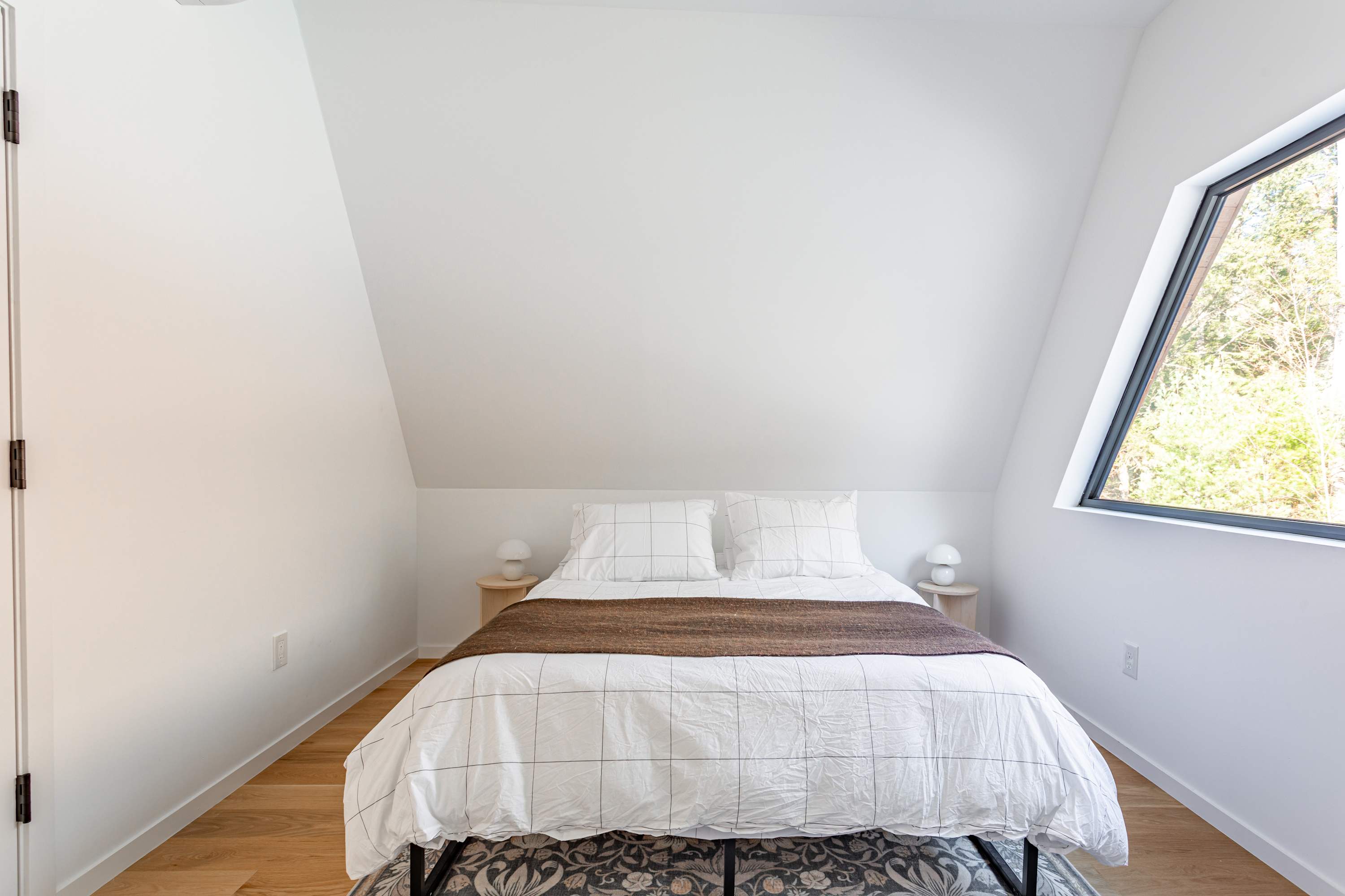 A cozy bedroom with white walls and a sloped ceiling, featuring a queen bed with white bedding and a brown throw. Two small round nightstands with lamps flank the bed, which rests on a patterned rug. A large angled window offers a view of the forest.