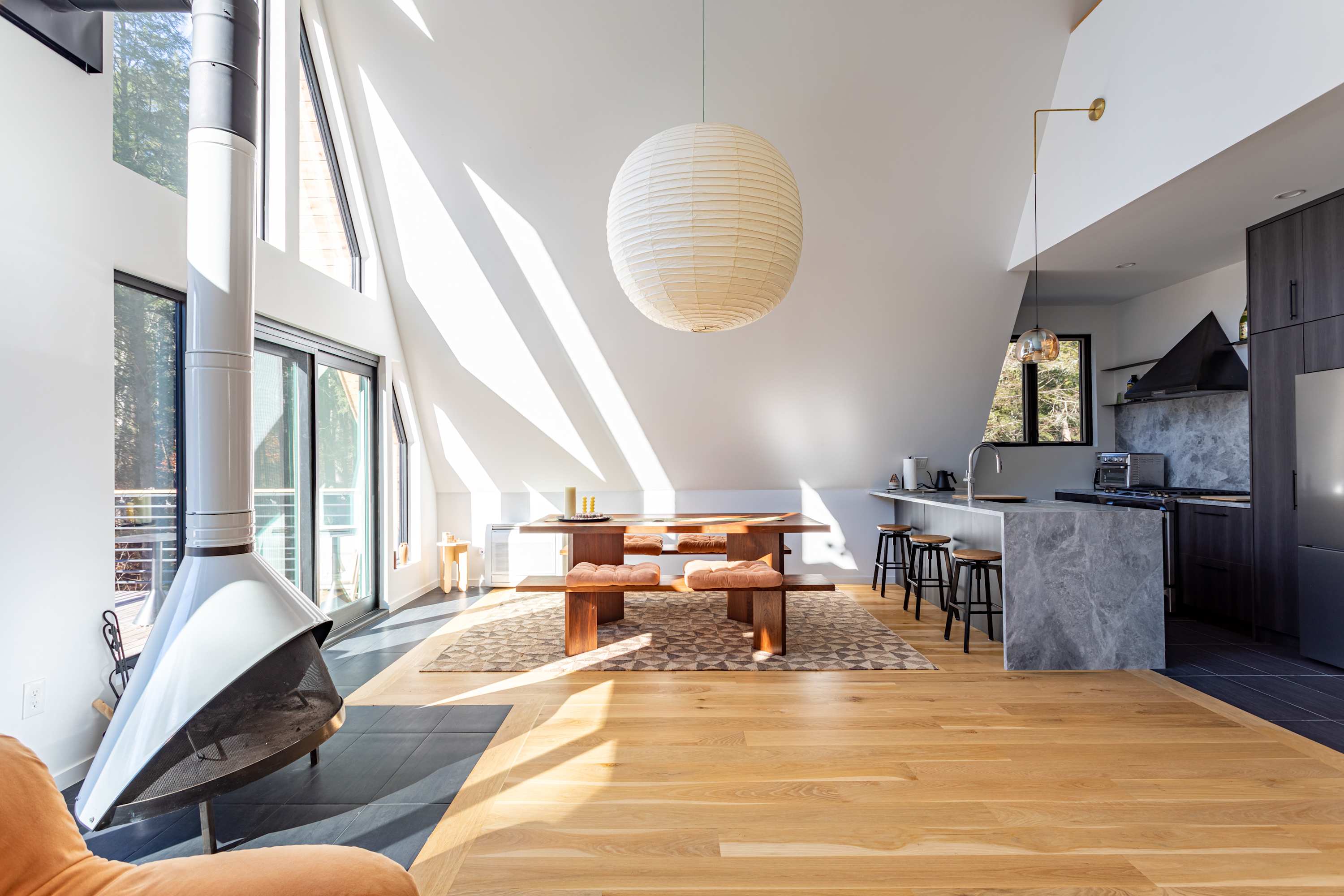 A bright, open-concept A-frame space with a wooden dining table and benches on a patterned rug, illuminated by a large paper lantern. A modern kitchen with dark cabinetry and a gray island is visible to the right, and a freestanding fireplace to the left.