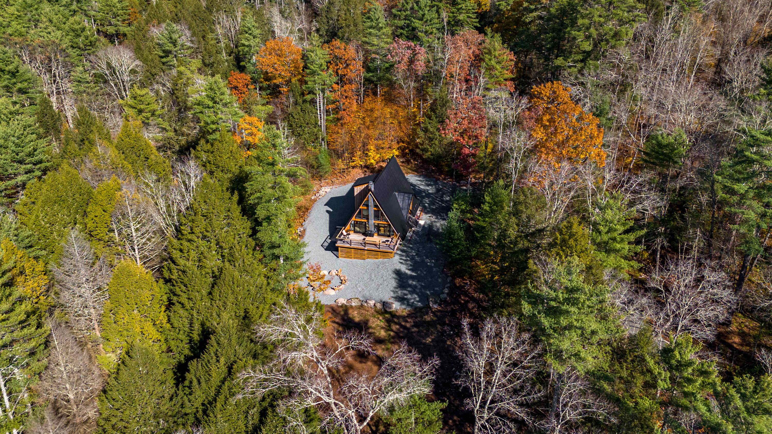 An aerial view showcases a modern black A-frame house with a wooden deck, nestled amidst a dense forest displaying vibrant autumn foliage and evergreen trees. A gravel driveway encircles the house, leading to a small fire pit area.