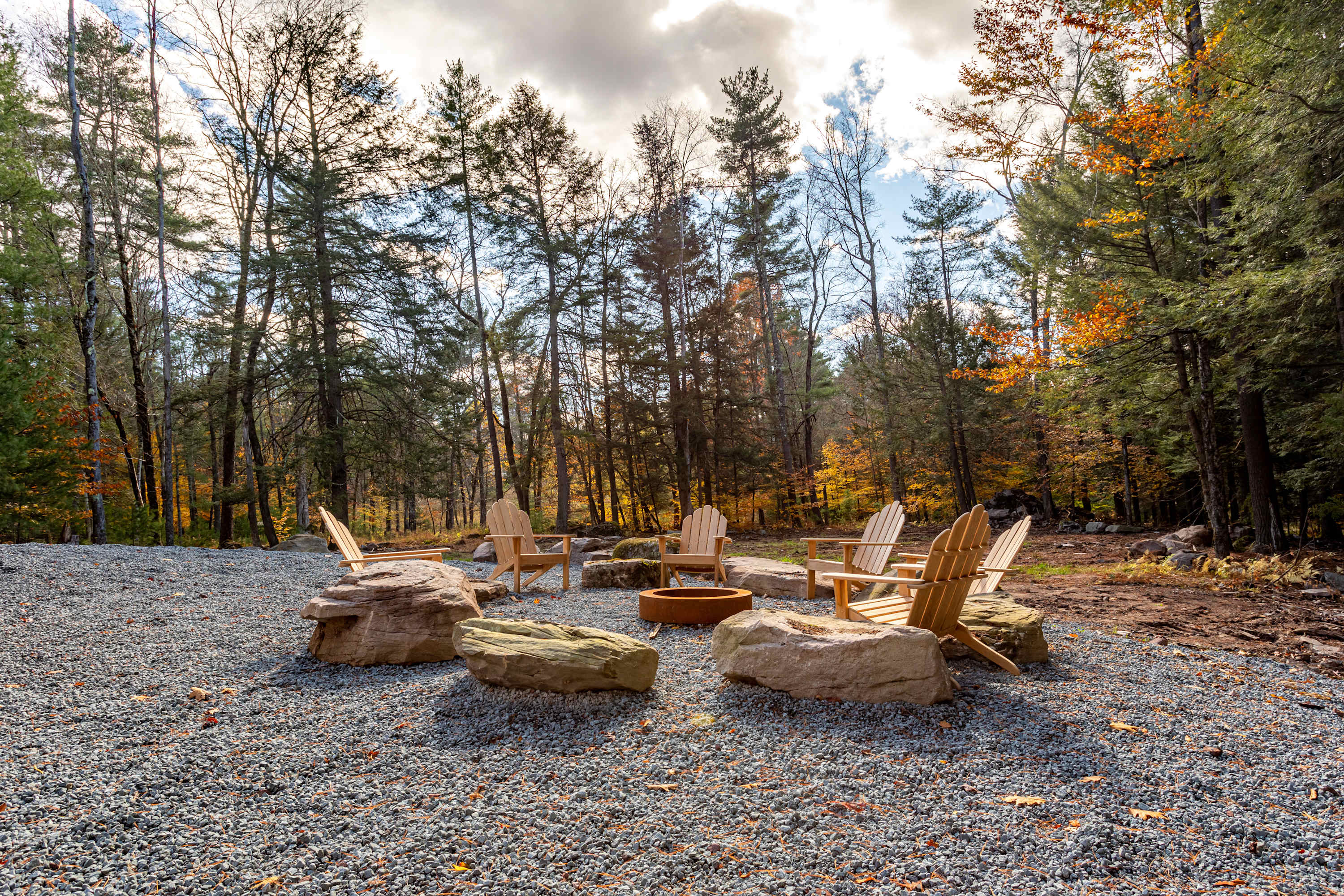 A circular fire pit area is set on a gravel ground, surrounded by large natural rocks and several wooden Adirondack chairs, with a dense forest backdrop under a cloudy sky.