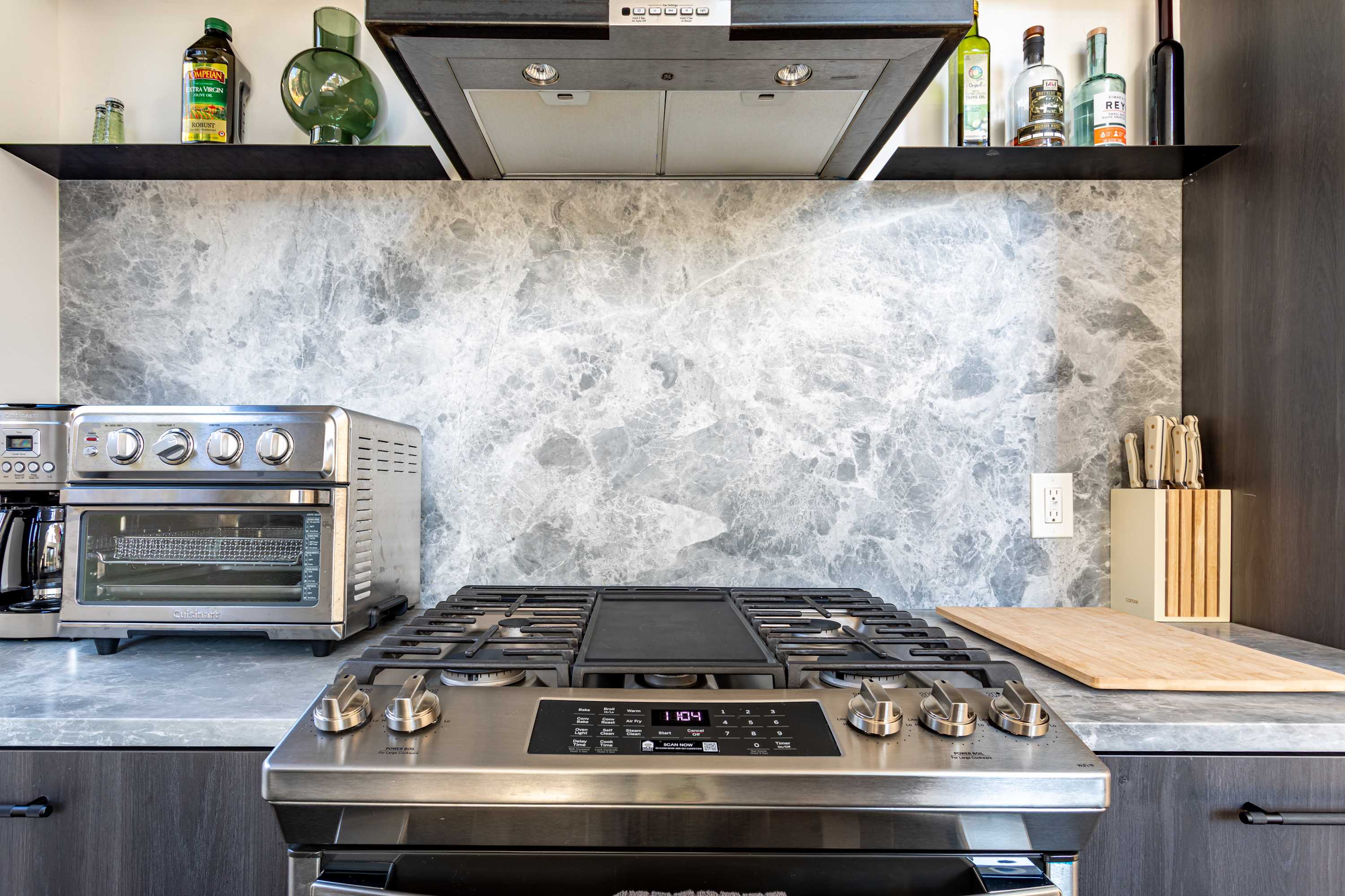 A detailed view of the kitchen's cooking area, featuring a stainless steel gas range with a griddle, a matching range hood, and a gray marble-look backsplash. Open shelving above holds decorative bottles.