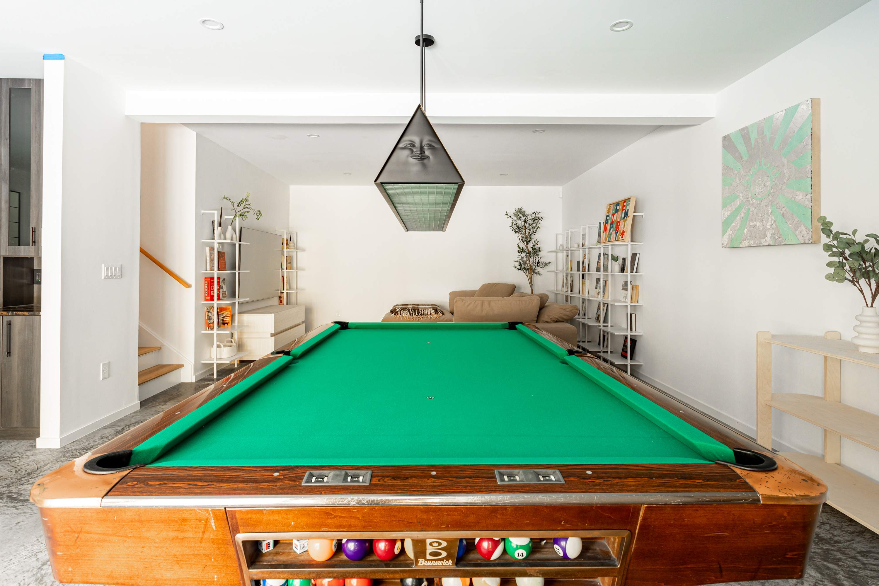 A game room with polished concrete floors and white walls, dominated by a classic wooden pool table with green felt under a unique black pendant light. White shelving units line the walls in the background, leading to a seating area.