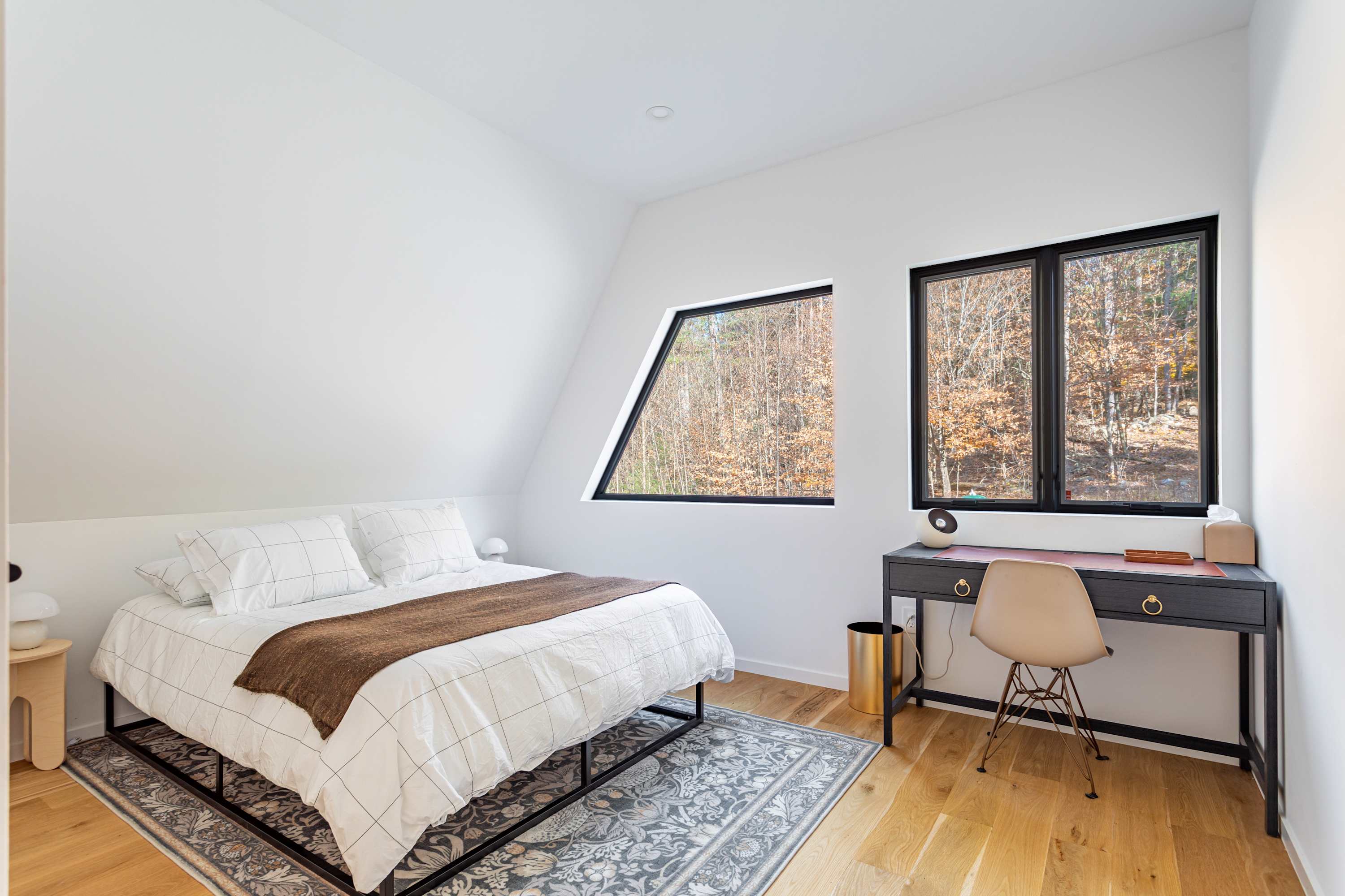 A bright bedroom with white walls, a sloped ceiling, and light wood floors. A queen bed with white bedding and a brown throw is on a patterned rug, facing two large windows with black frames overlooking a forest. A dark desk with a light chair is to the right.