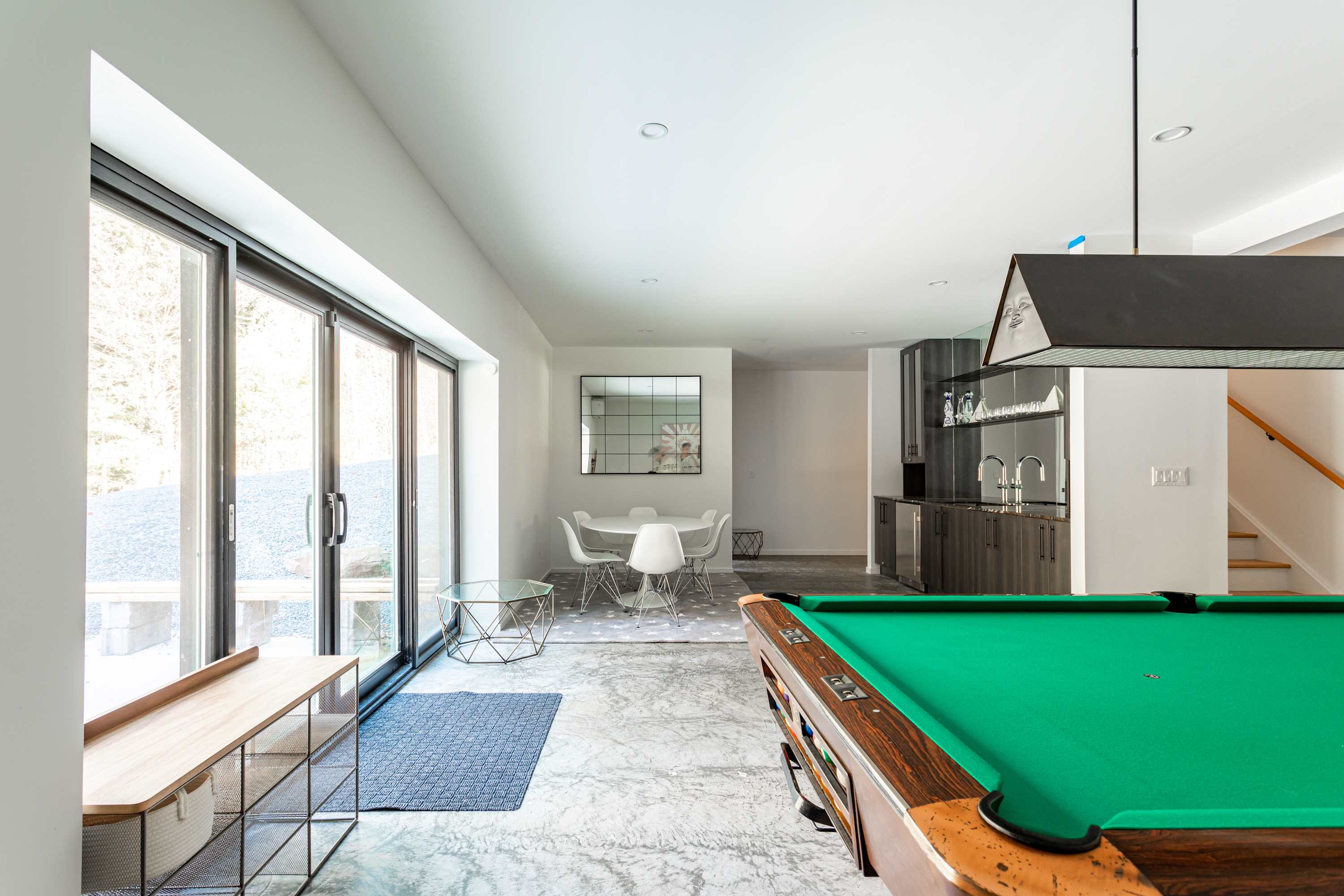 A bright game room and dining area with polished concrete floors and white walls. A pool table is in the foreground, leading to a dining space with a white round table and chairs. Large sliding glass doors open to the outside, and a wet bar area is visible to the right.
