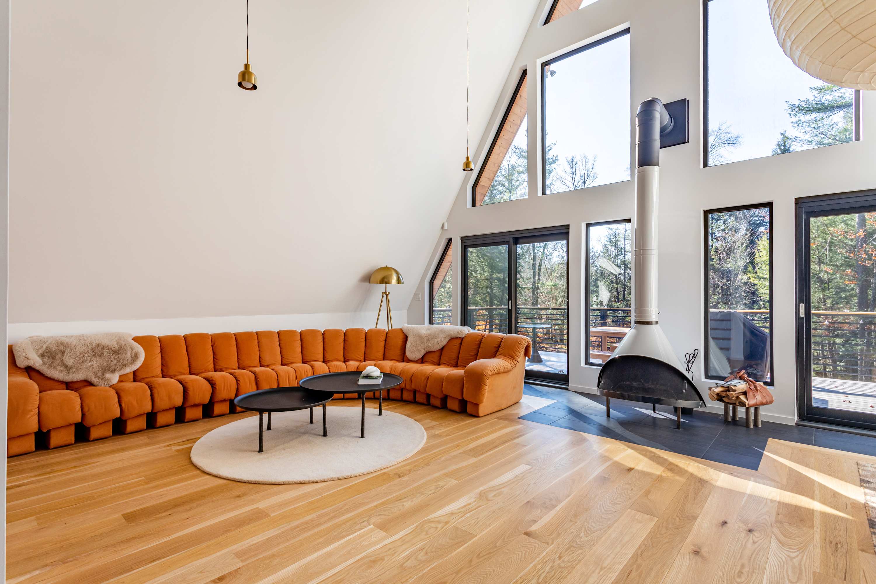 A modern A-frame living room features an orange modular sofa, two black round coffee tables on a white rug, and a freestanding black and white fireplace. Large windows offer views of the surrounding trees.