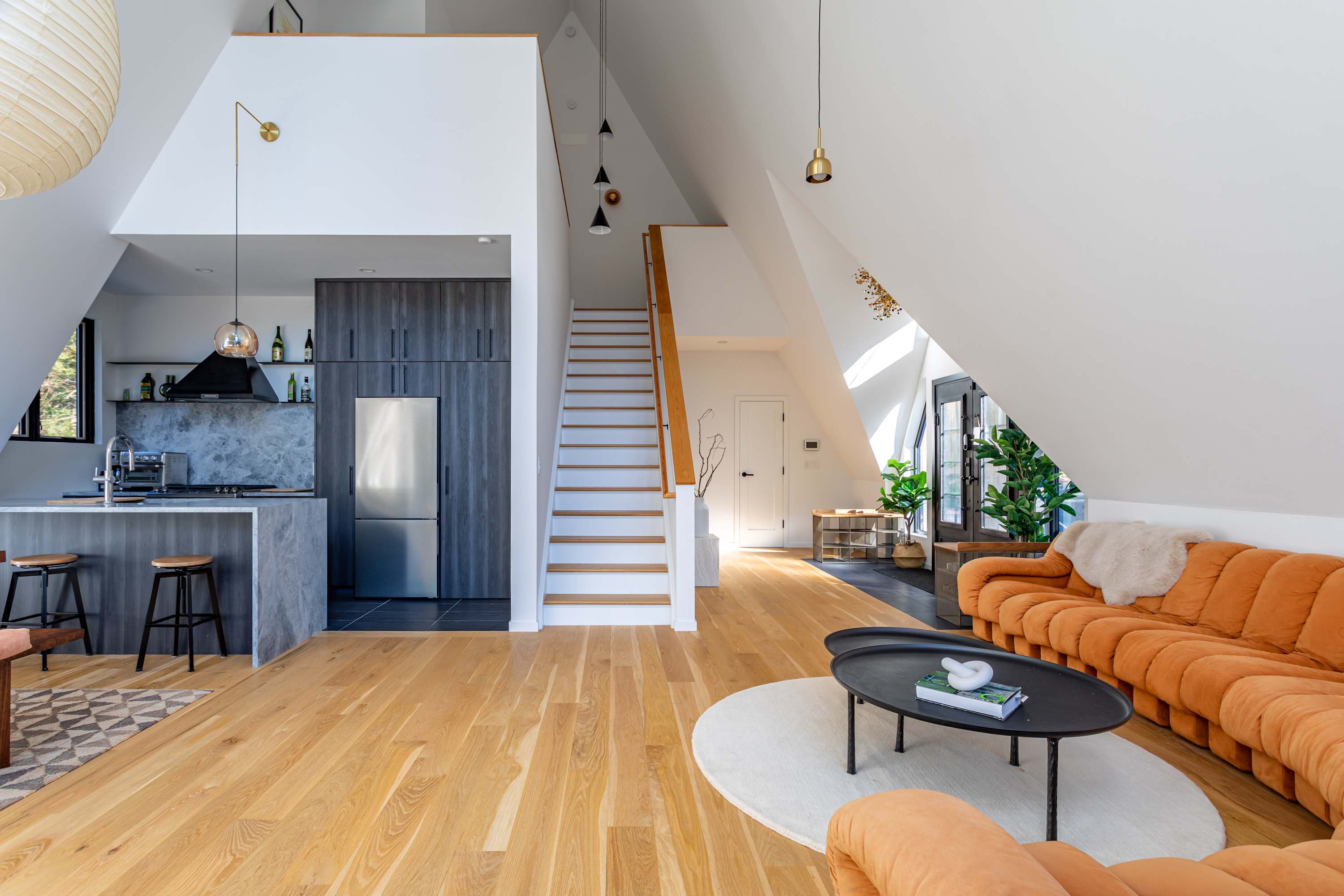 An open-concept living area with light hardwood floors and a high, sloped white ceiling. An orange sectional sofa faces a black coffee table on a round rug. In the background, a modern kitchen with dark cabinetry, a gray island, and stainless steel appliances is visible. A staircase leads upstairs.