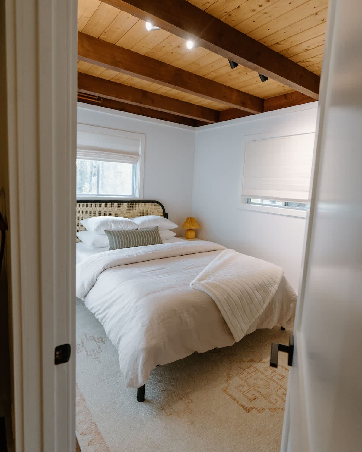 A cozy bedroom with a light wood slatted headboard bed, white bedding, and a striped accent pillow. The room features a rustic wooden beam ceiling, white walls, and a patterned rug.