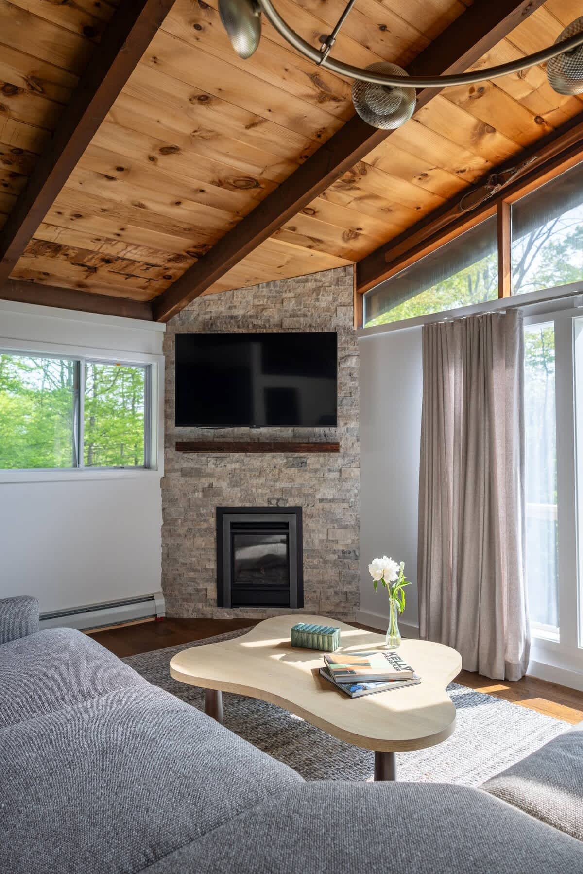 A cozy living room boasts a grey sectional sofa, a unique organic-shaped coffee table, and a stone-clad fireplace with a mounted TV. Exposed wooden beams on the ceiling add rustic charm, complemented by large windows.