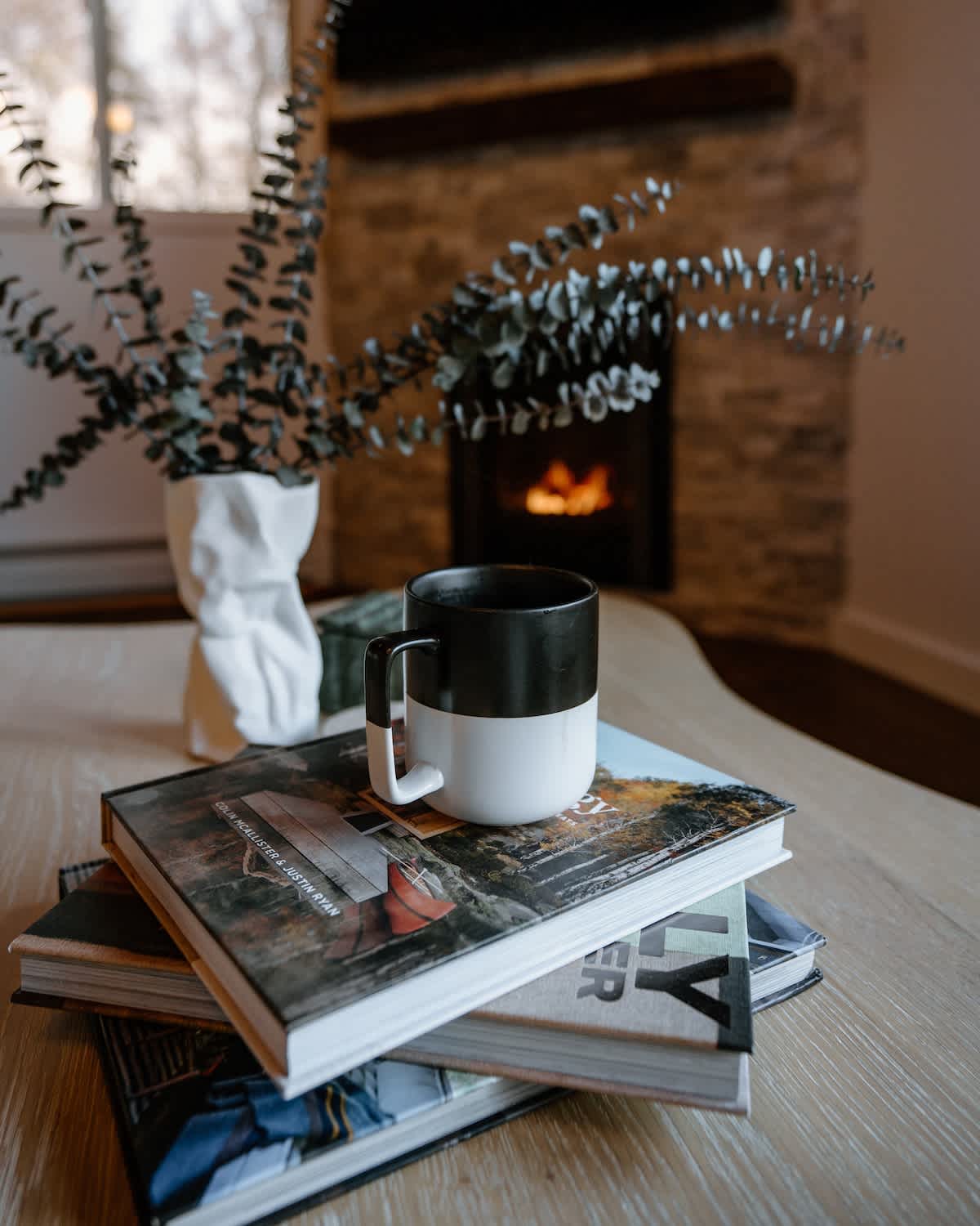 A close-up of a light wood coffee table with a stack of books and a two-toned black and white mug. In the background, a fireplace with a warm fire and eucalyptus decor are visible.