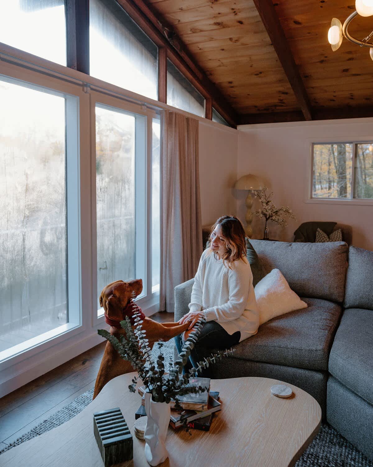 A bright living room features a large grey sectional sofa, a light wood coffee table with eucalyptus decor, and expansive windows looking out to frosted trees. A woman and a Vizsla dog are interacting by the window.
