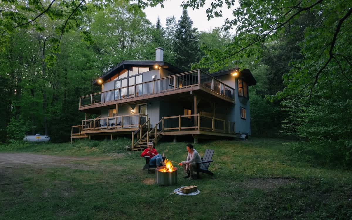 Exterior view of a modern chalet-style house with multiple decks, set amidst a lush green forest, with two people enjoying a fire pit in the foreground.