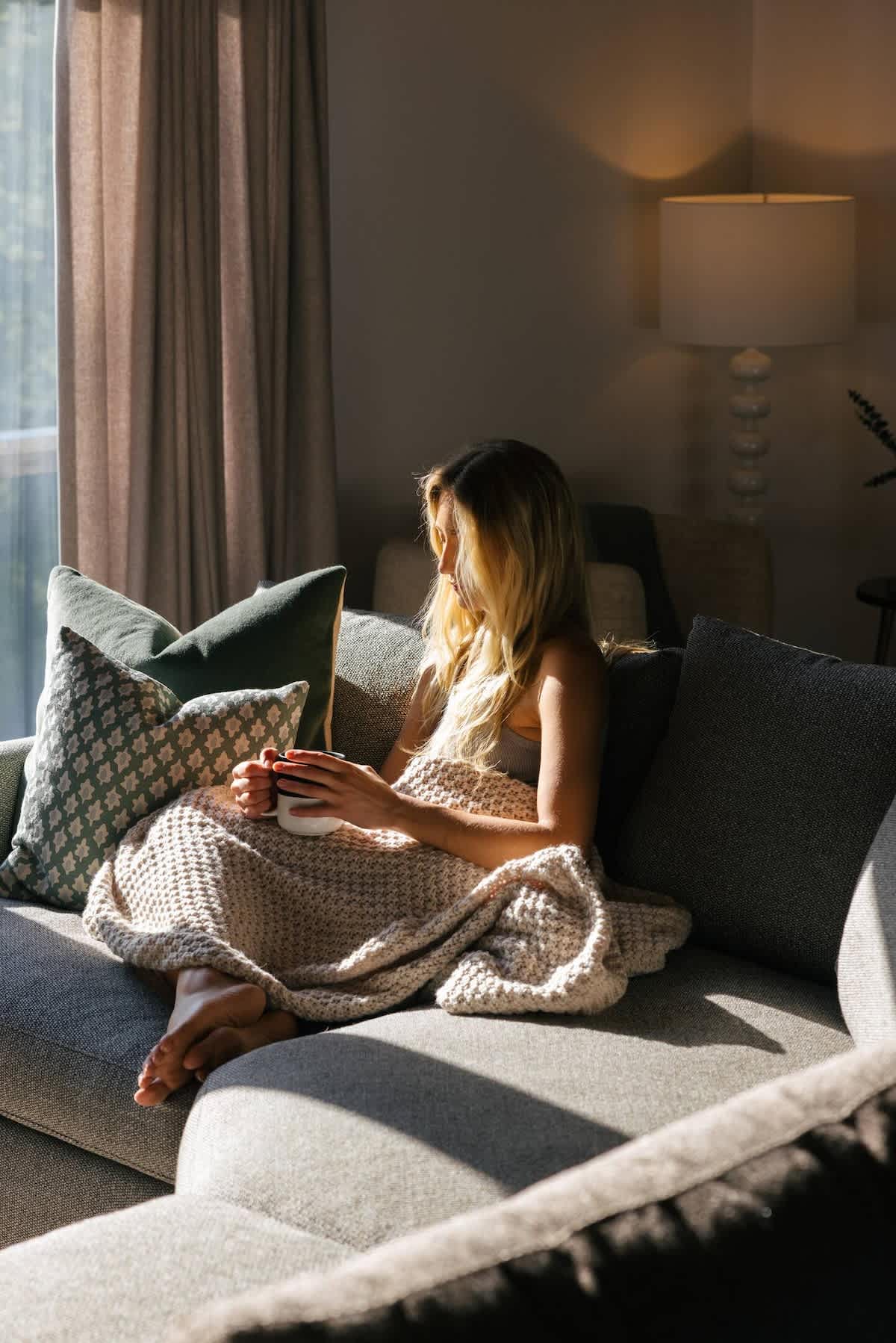 A person is comfortably curled up on a grey sofa with a knitted blanket and a mug, bathed in warm sunlight streaming through a large window. Decorative pillows and a floor lamp complete the inviting scene.
