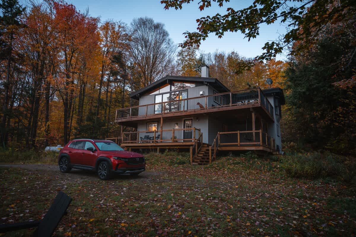 The modern chalet, with its multi-level deck and large windows, is nestled among vibrant autumn trees. A red SUV is parked in the gravel driveway, surrounded by fallen leaves.