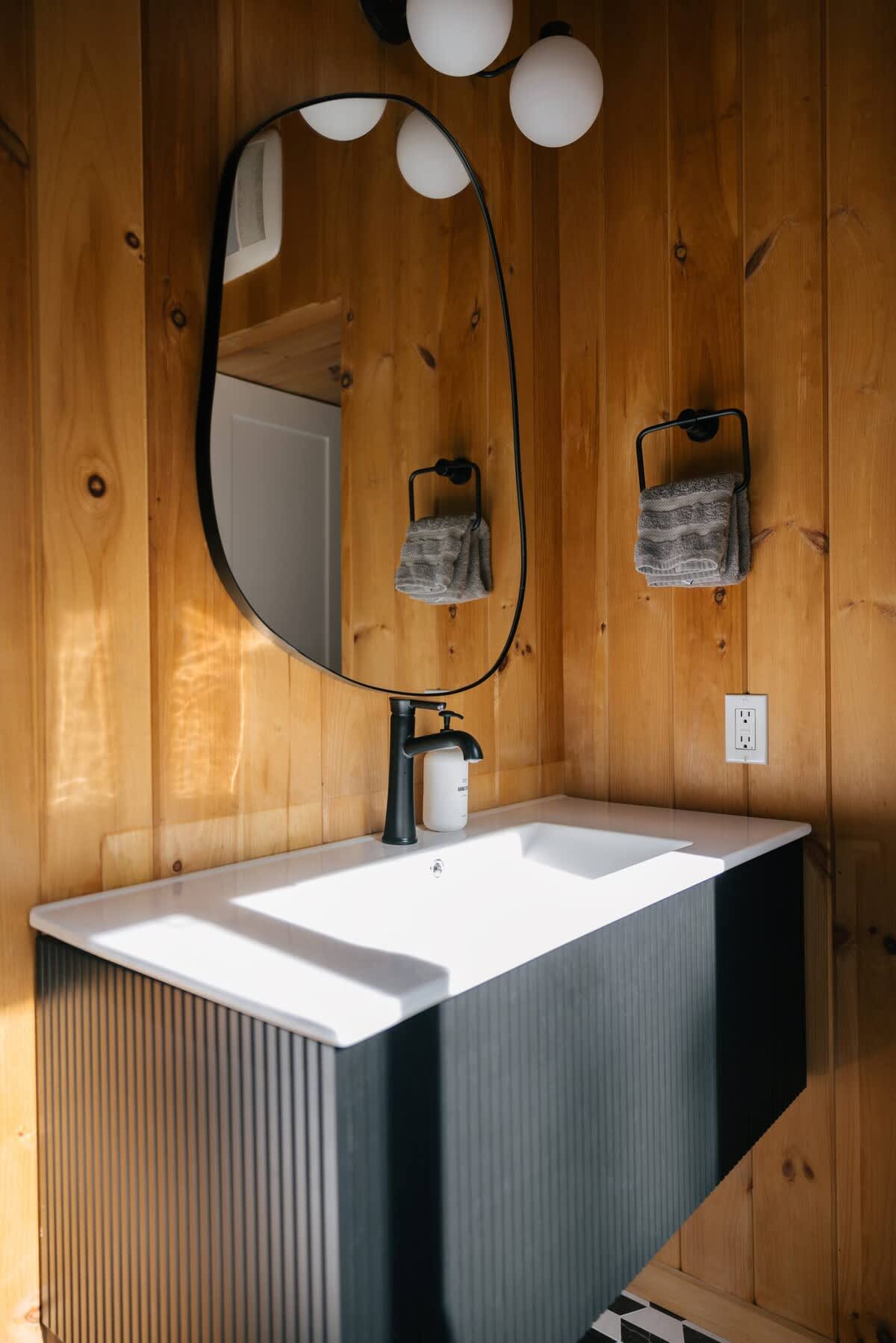 Modern bathroom with warm wood paneling, a unique organic-shaped black mirror, and a dark fluted vanity with a white integrated sink and black faucet.