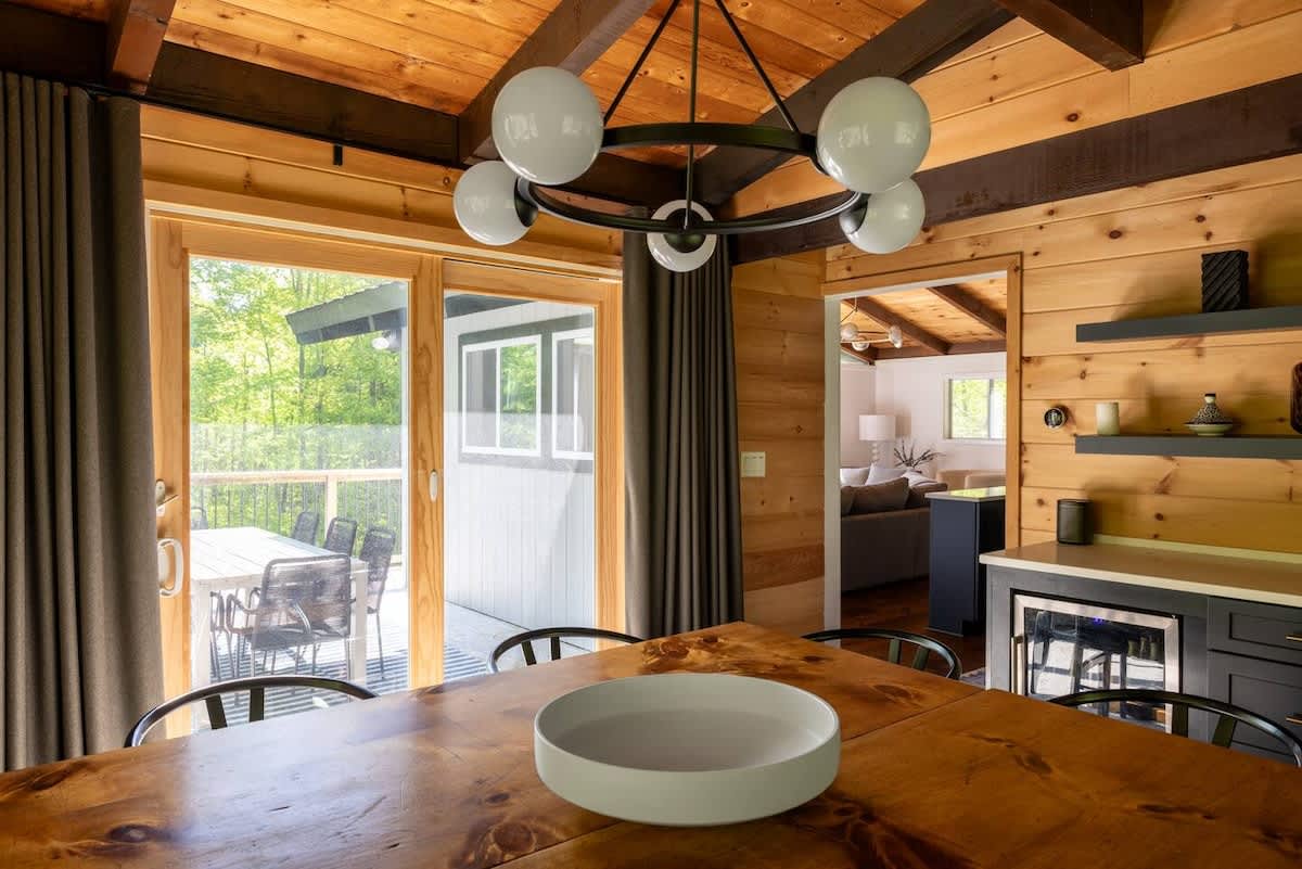 A rustic dining room with wood-paneled walls and a large wooden table opens onto an outdoor deck through sliding glass doors. A modern chandelier and a built-in bar area with a beverage fridge are visible.