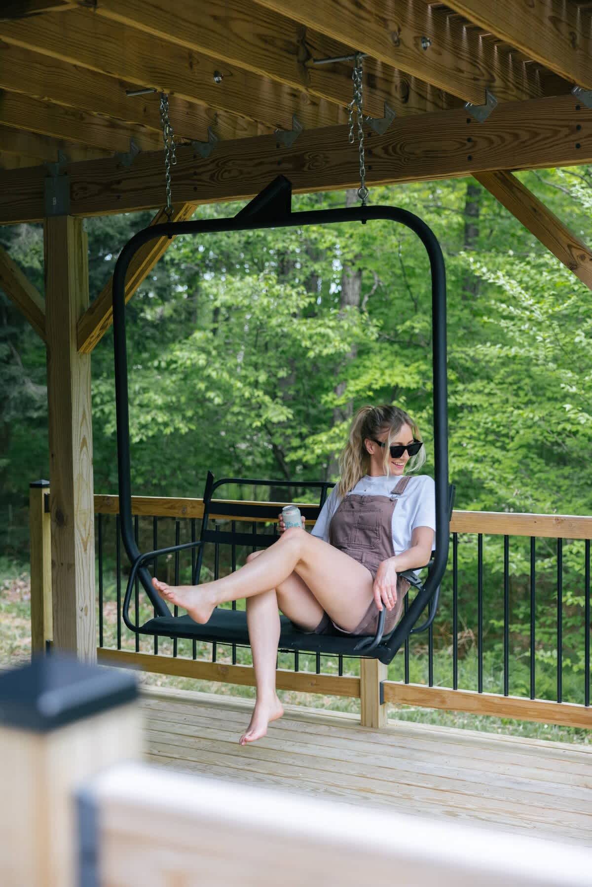 A person relaxes on a unique black ski lift chair swing, suspended from the wooden deck ceiling, enjoying the lush green forest view. The chair is made of metal and black seating material.