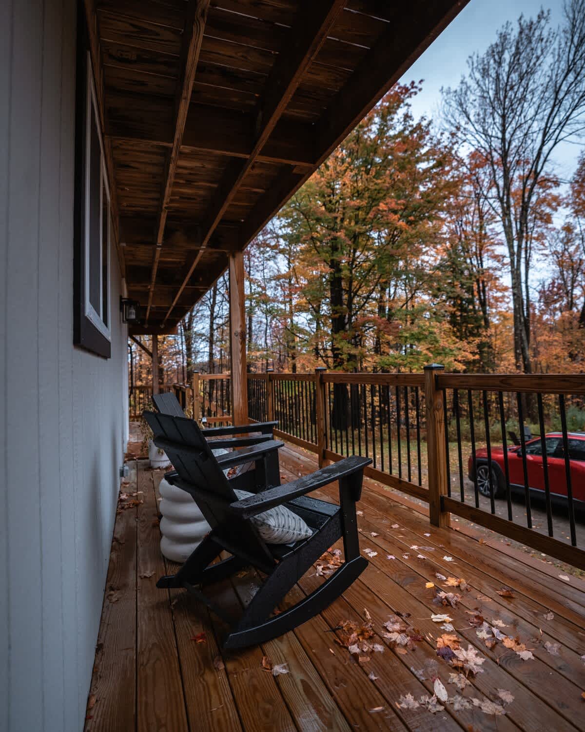 Covered wooden deck with black rocking chairs and a side table, surrounded by autumn trees, offering a cozy outdoor seating area.