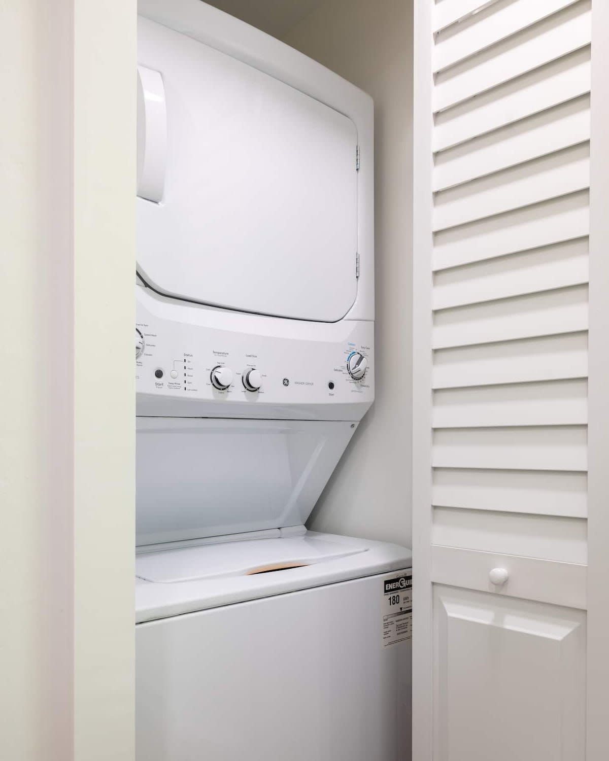 A compact laundry closet features a white stacked washer and dryer unit, neatly tucked behind louvered bi-fold doors. The space is clean and functional, with a visible energy guide sticker on the lower appliance.