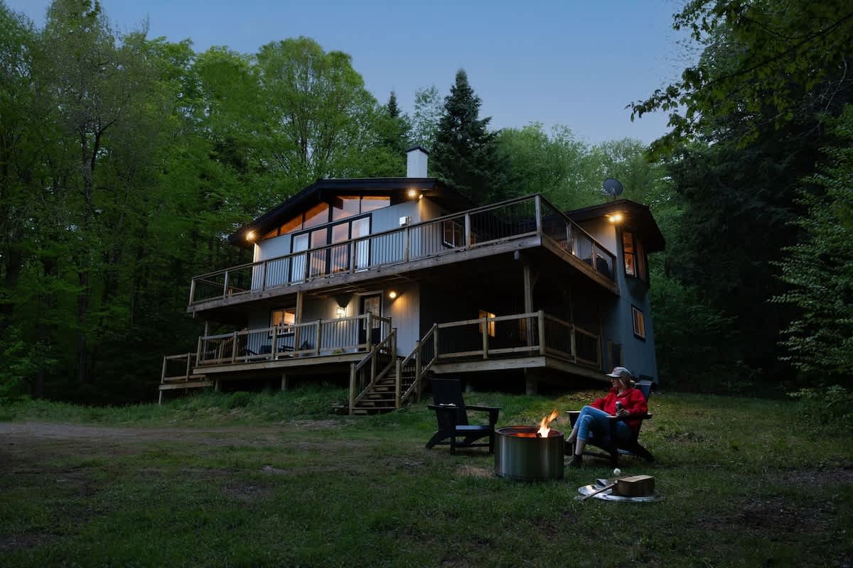 The modern chalet is illuminated at dusk, surrounded by lush trees, with a person relaxing by a glowing fire pit in the grassy yard. Multiple decks and outdoor seating areas are visible.