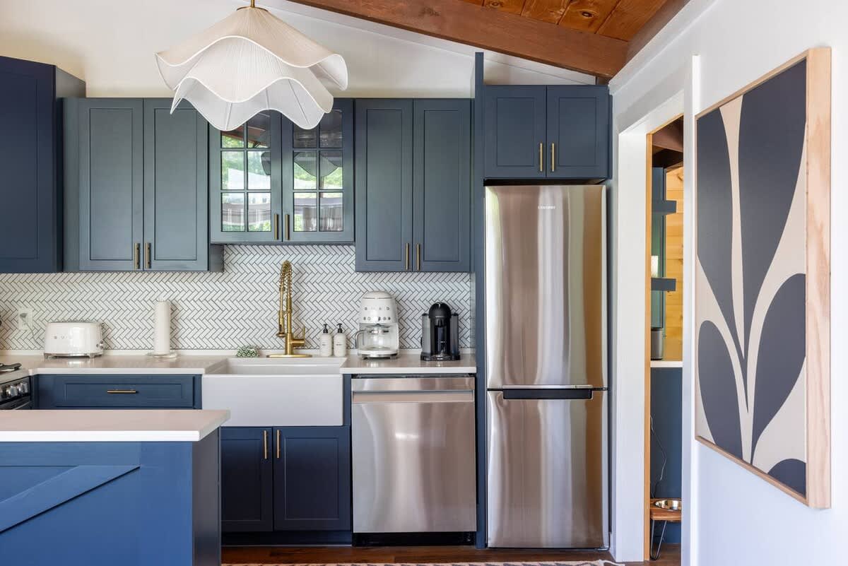 A bright kitchen showcases navy blue shaker cabinets with gold pulls, white countertops, and a herringbone tile backsplash. Stainless steel appliances, including a French door refrigerator and dishwasher, are visible alongside a unique white pendant light.