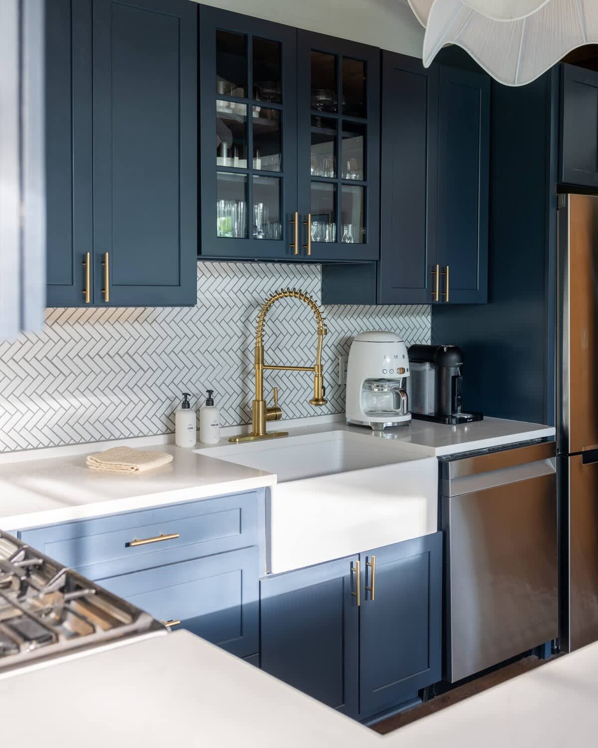 Modern kitchen with dark blue shaker cabinets, white quartz countertops, and a white herringbone subway tile backsplash. A gold pull-down faucet stands out against the white farmhouse sink, with stainless steel appliances visible.