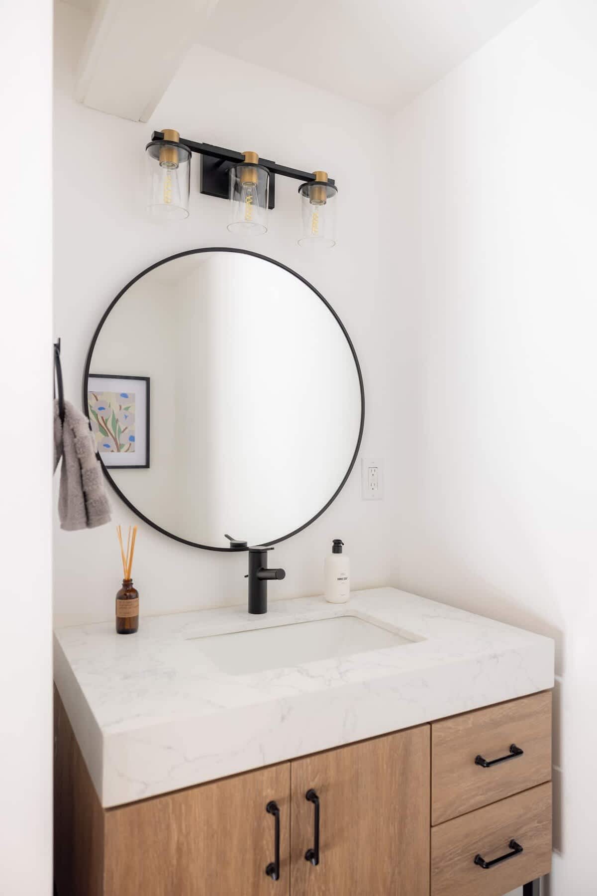 Bright modern bathroom with white walls, a round black-framed mirror, a light wood vanity with a white marble-patterned countertop, and a black faucet.
