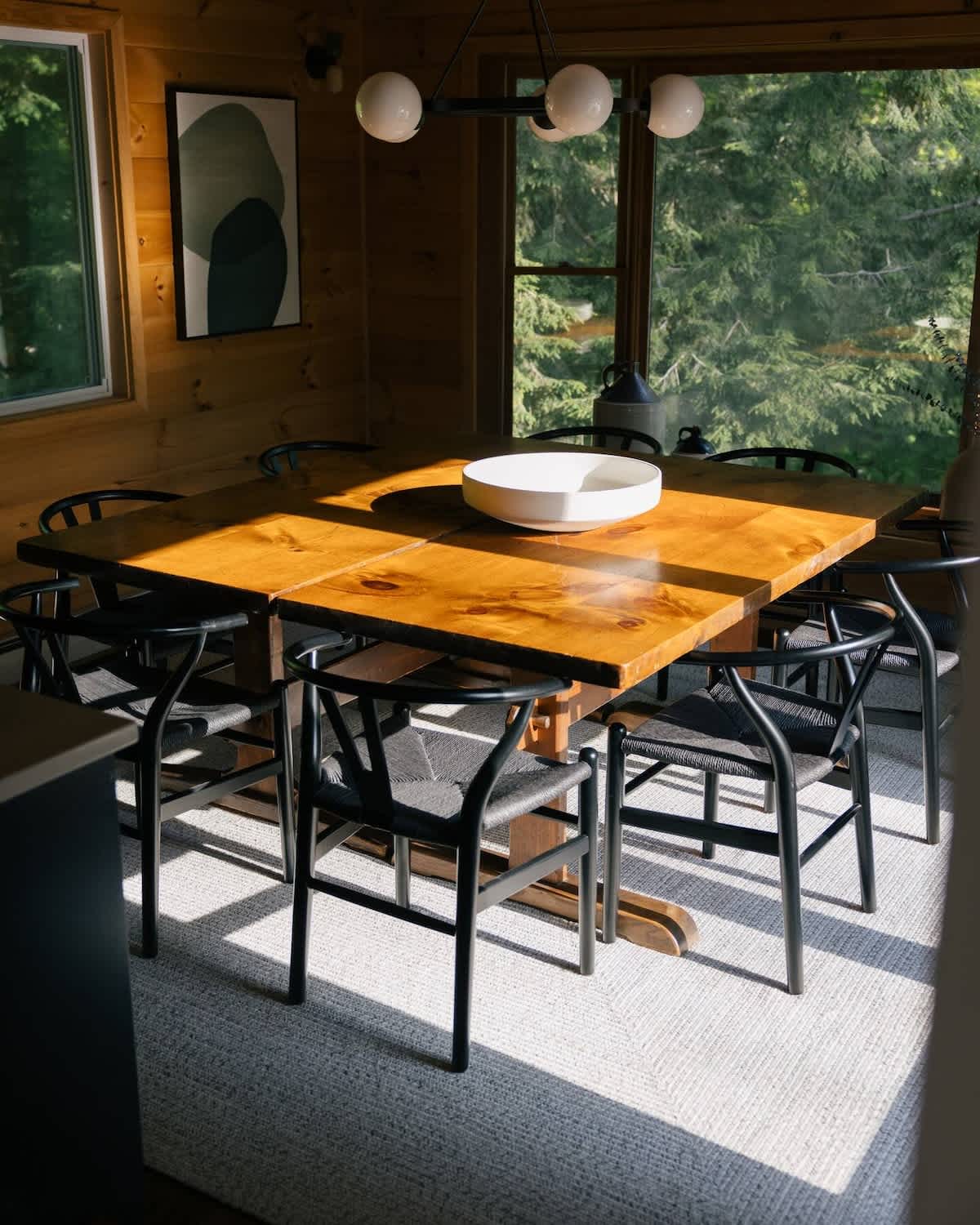 A sunlit dining room features a large wooden table with black wishbone chairs, set on a light textured rug, with warm wood paneling and large windows overlooking green trees. A modern chandelier hangs overhead.