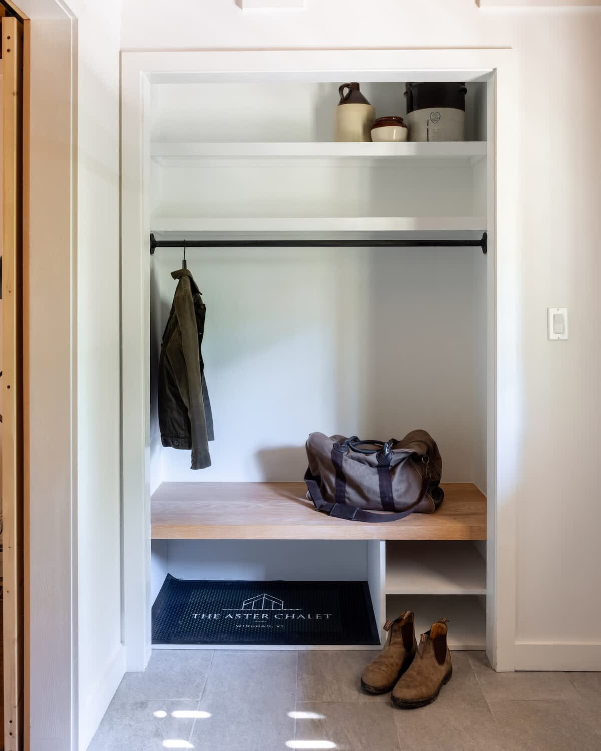 A bright, minimalist mudroom closet features white shelving, a wooden bench, and a black hanging rod with a jacket. A duffel bag and boots rest on the bench and floor, with a welcome mat visible.