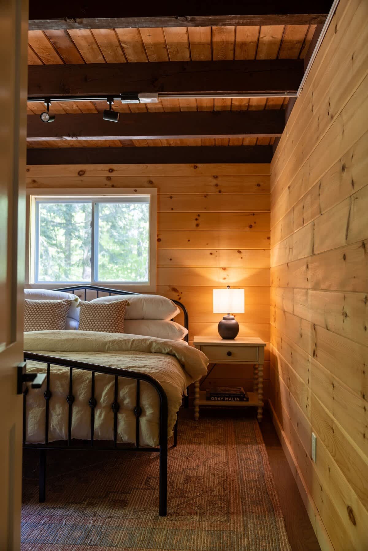 Cozy bedroom featuring warm wood-paneled walls and ceiling with exposed dark beams. A black metal bed frame with light bedding is positioned next to a window overlooking greenery, with a small nightstand and lamp.