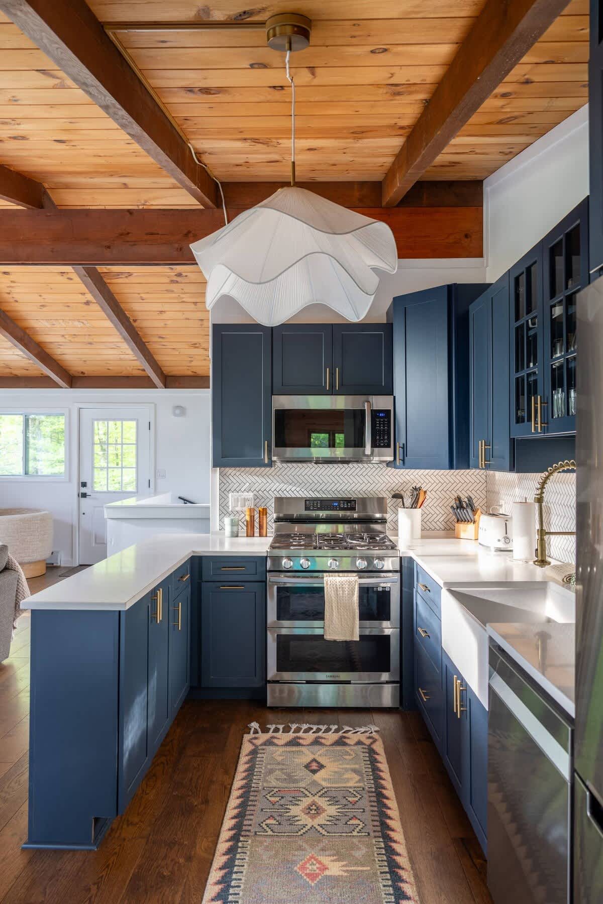 U-shaped kitchen featuring dark blue shaker cabinets, white countertops, and a herringbone tile backsplash, with stainless steel appliances including a gas range. A large white pendant light hangs from the wood-paneled ceiling with exposed beams.