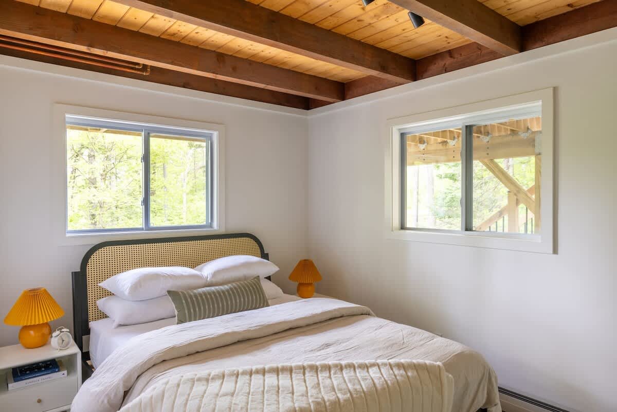 Serene bedroom with white walls and a light wood and cane headboard, featuring a bed with white bedding, two white nightstands with unique orange lamps, and two windows.