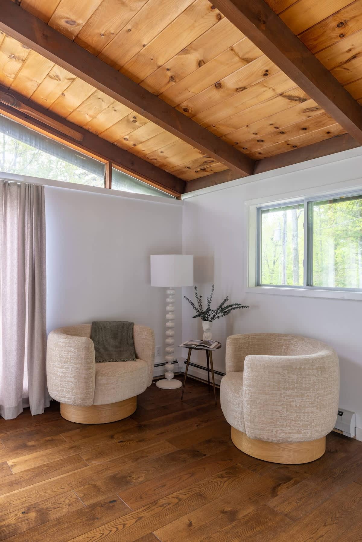 Bright sitting area with two rounded cream-colored armchairs and a tall white floor lamp, set on a dark wood floor. The room features a vaulted wood-paneled ceiling with dark beams and large windows.