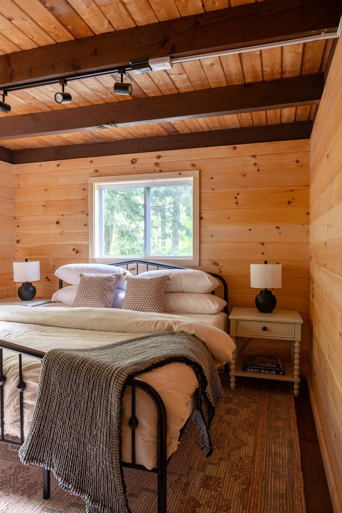 Cozy bedroom with warm wood paneling and exposed ceiling beams, featuring a black metal bed frame with white bedding, two nightstands with lamps, and a window overlooking trees.