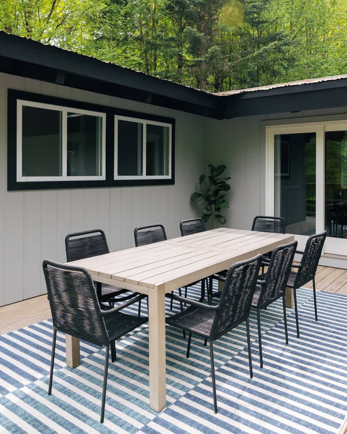 An outdoor dining area on a wooden deck features a light-colored rectangular dining table surrounded by eight dark woven chairs, set atop a blue and white striped outdoor rug. The area is surrounded by lush trees.