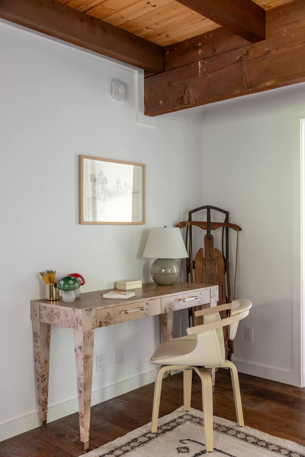 Bright workspace with a light-colored patterned desk and a modern cream-colored chair, set against a white wall with a framed picture. Dark exposed beams contrast with the white ceiling, and a vintage sled leans in the corner.