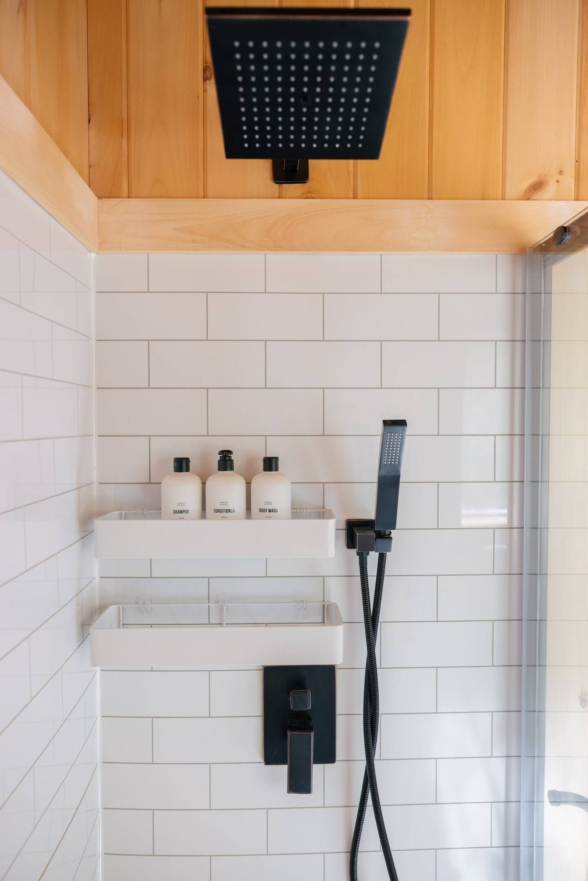 Modern shower with white subway tiles, a black square rain shower head, and a black handheld shower. Three white bottles labeled "Shampoo," "Conditioner," and "Body Wash" are neatly arranged on a white shelf.