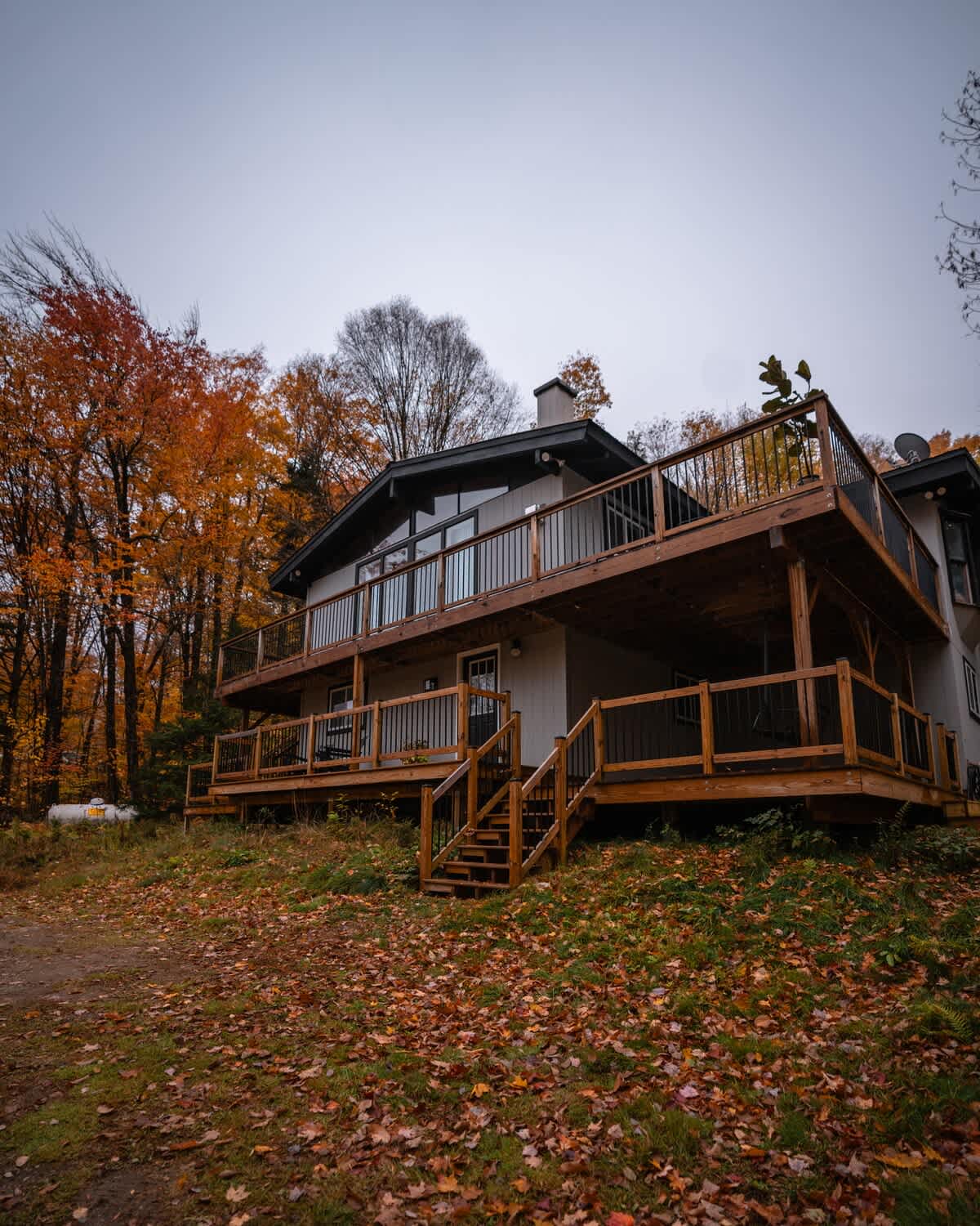 Exterior view of a multi-level chalet-style house with extensive wooden decks and railings, surrounded by colorful autumn trees under an overcast sky.