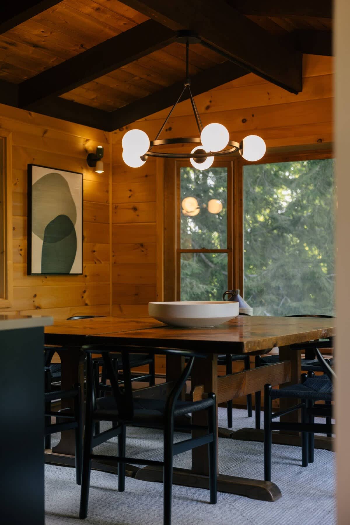 Rustic dining area featuring a large wooden table surrounded by black chairs, under a modern black chandelier with globe lights. Wood-paneled walls and ceiling with dark beams create a warm ambiance, with a large window offering outdoor views.