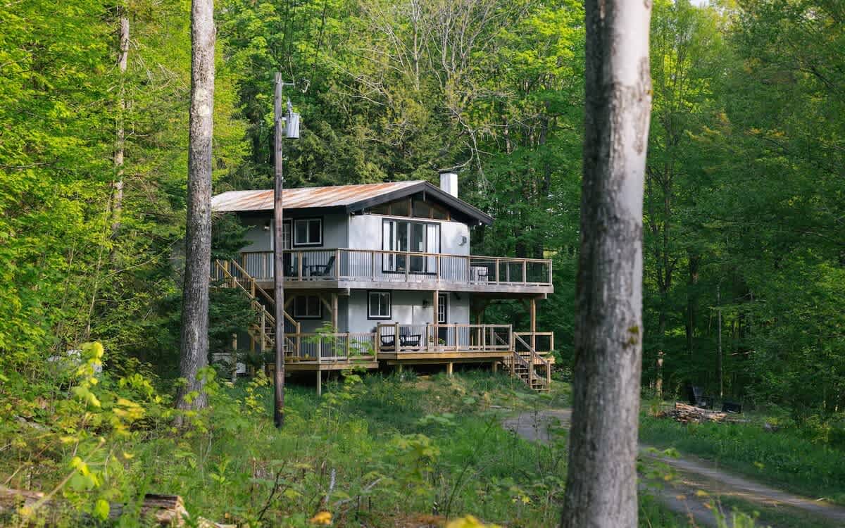 A modern chalet-style house with grey siding and a rustic metal roof is nestled among dense green trees, featuring multiple levels of wooden decks and railings. A dirt path leads up to the secluded forest retreat.