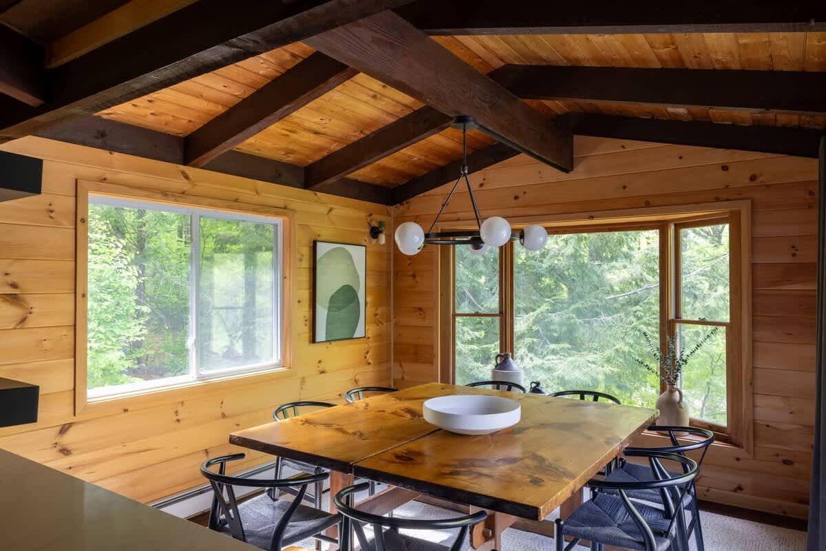 Rustic dining room with extensive wood paneling and exposed ceiling beams, featuring a large square wooden table surrounded by black chairs, and large windows overlooking a forest.