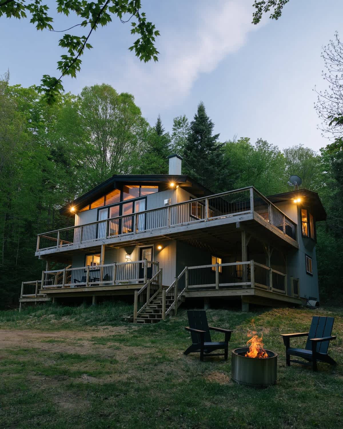 Charming two-story chalet with multiple decks and large windows, nestled among lush green trees under a twilight sky. A fire pit with two Adirondack chairs sits in the grassy yard, illuminated by the house's warm lights.