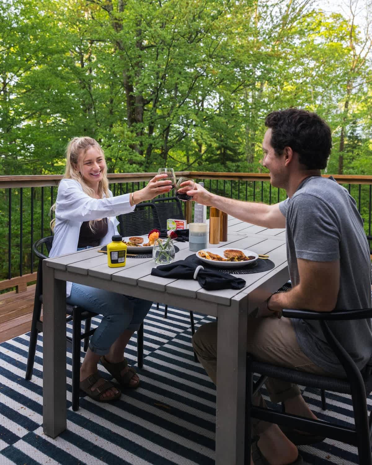 A couple enjoys an outdoor meal on a spacious wooden deck, seated at a modern grey dining table with black chairs over a striped rug. Lush green trees provide a natural backdrop, creating a serene dining experience.