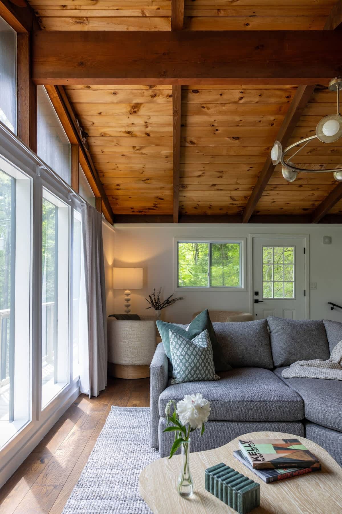 Spacious living room with a large grey sectional sofa and a light wood coffee table, featuring a vaulted wood-paneled ceiling with dark beams. Floor-to-ceiling windows offer views of the surrounding trees, and a modern chandelier hangs overhead.