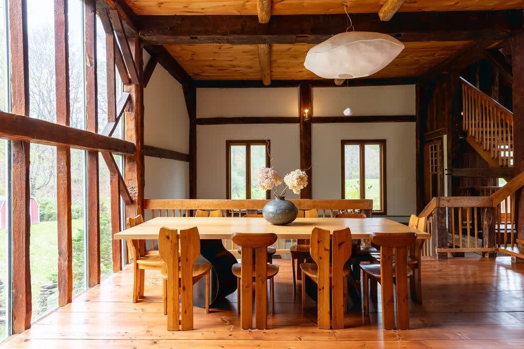 Spacious dining area with a long wooden table and matching chairs, including a bench, set on warm hardwood floors. Large windows provide ample natural light and views of the outdoors, with exposed wooden beams overhead and a unique white pendant light.