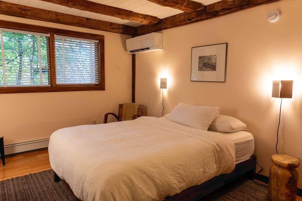 A cozy bedroom featuring a queen-sized bed with white bedding, light-colored walls, two wall-mounted lamps, and a window with blinds, all under a ceiling with exposed wooden beams.