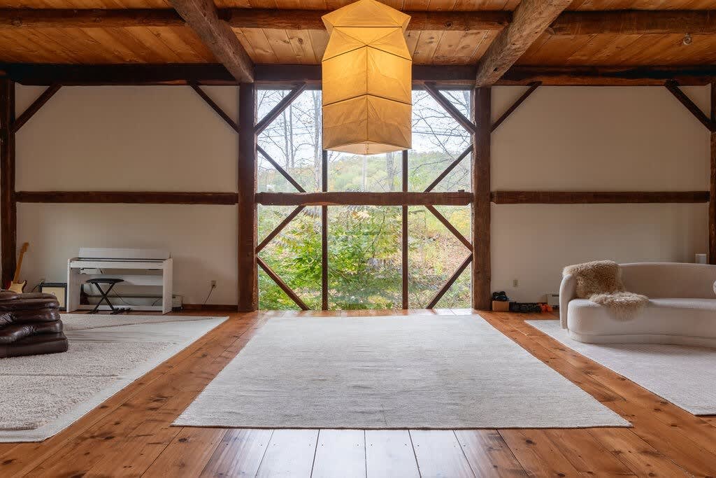 Expansive open-concept room with warm hardwood floors and multiple light area rugs, featuring a large geometric pendant light hanging from the high, exposed beam ceiling. A wall of windows offers views of the outdoors, with a piano and a curved sofa visible in the background.